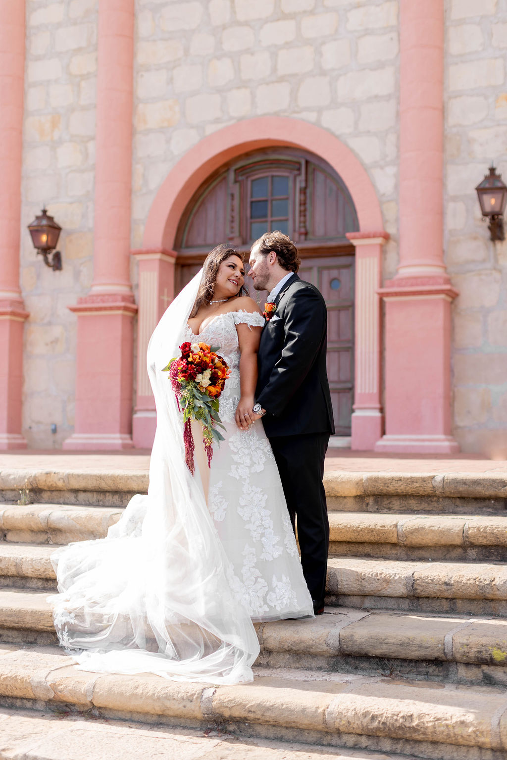 Bride and groom standing on the front steps of the Spanish Colonial style mission with pink pillars at Mission Santa Barbara wedding | Photo by Sarah Block Photography