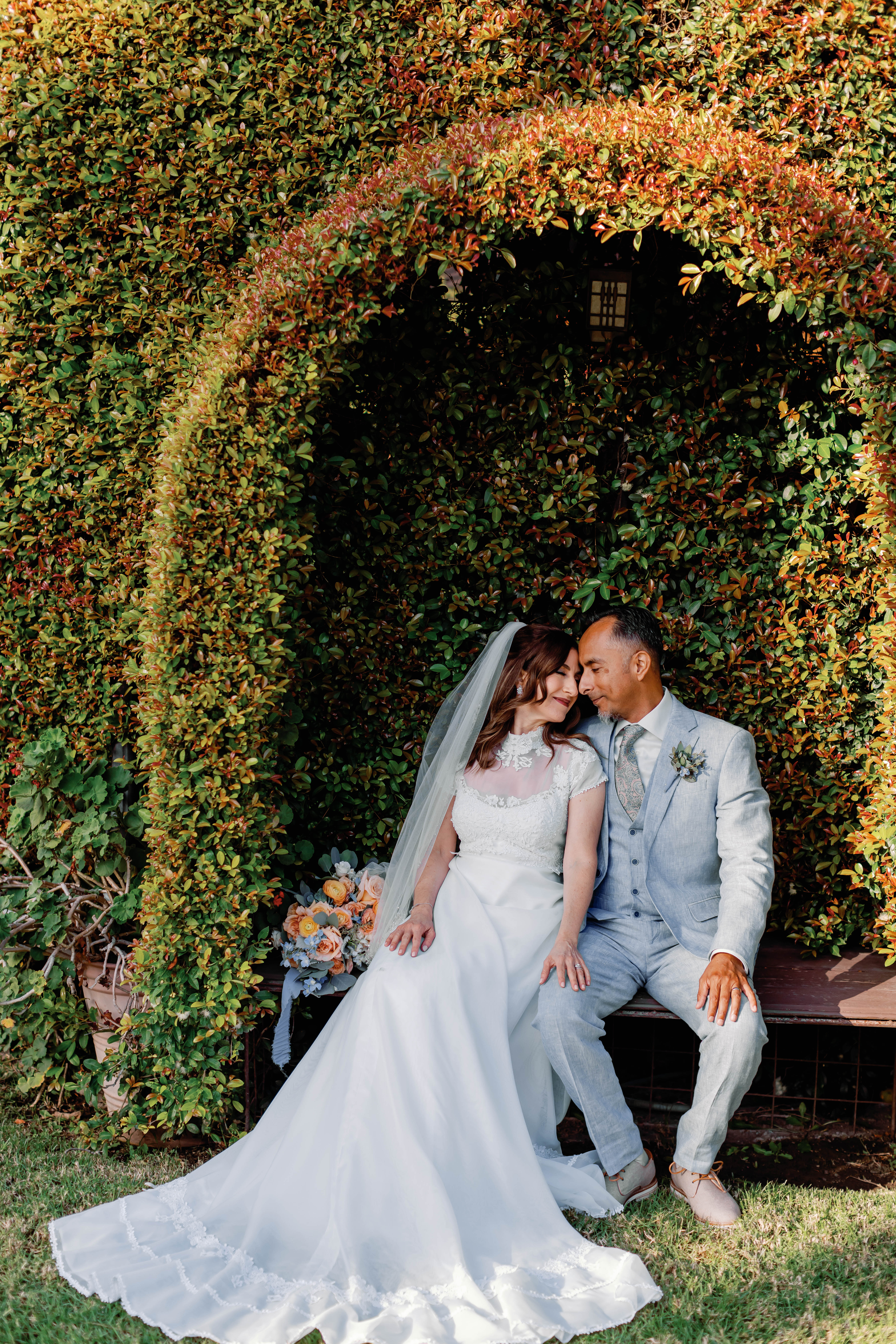 Bride and groom in garden under arbor sitting on a bench with wedding bouquet | Photo by Sarah Block Photography