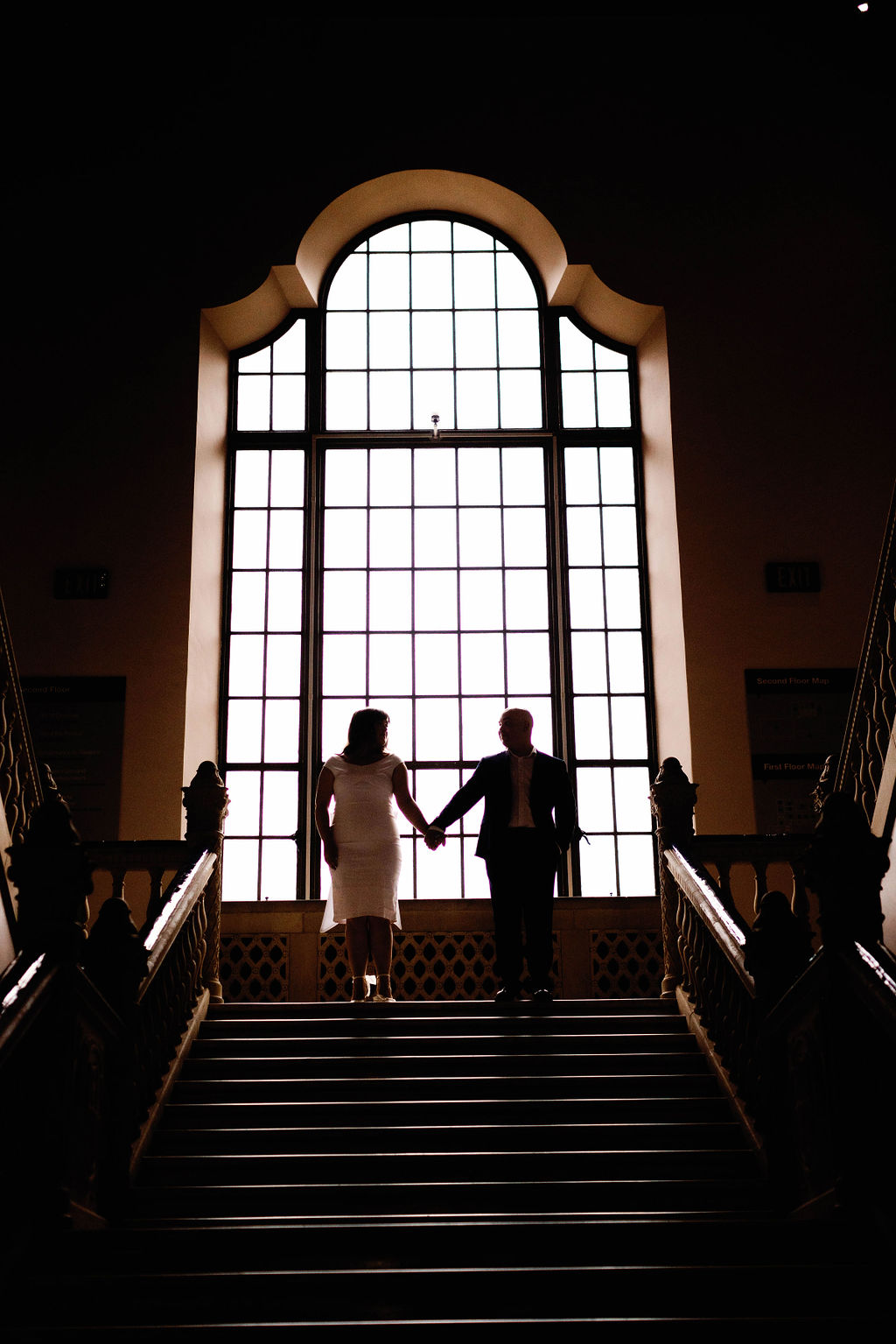 Jimmy and Hannah holding hands and walking down grand stairs with large window behind them. | Photo by California Photographer Sarah Block Photography