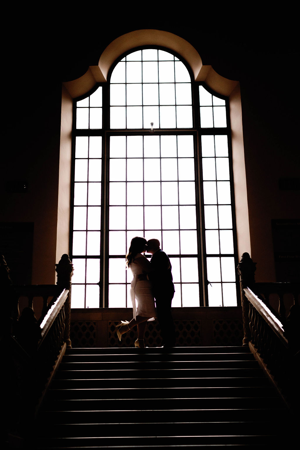 Hannah and Jimmy kissing on the stairs with the grand window at the San Diego Museum of Art. |Photo by California Photographer Sarah Block Photography