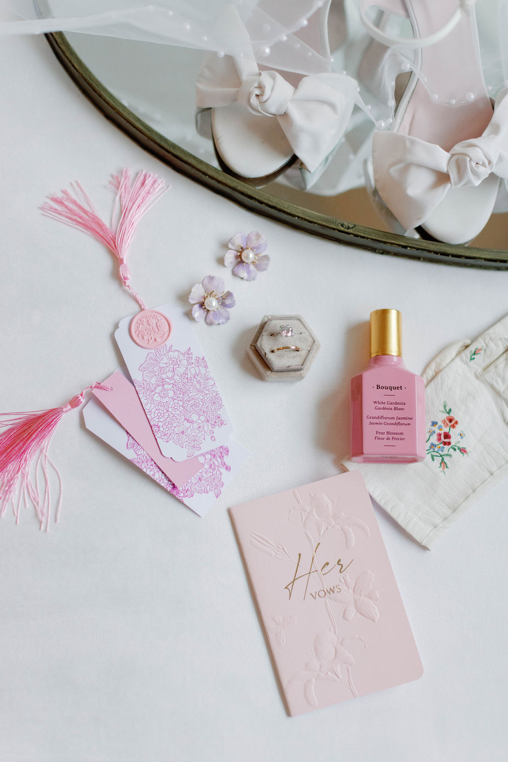 A photo of small wedding details including pink tassel and wax seal book marks, perfume, wedding rings, heirloom gloves , white wedding shoes with bows and earrings along with Vow books for a wedding hosted at The Grand Lady in Austin,Texas | Photo by Sarah Block of Sarah Block Photography.