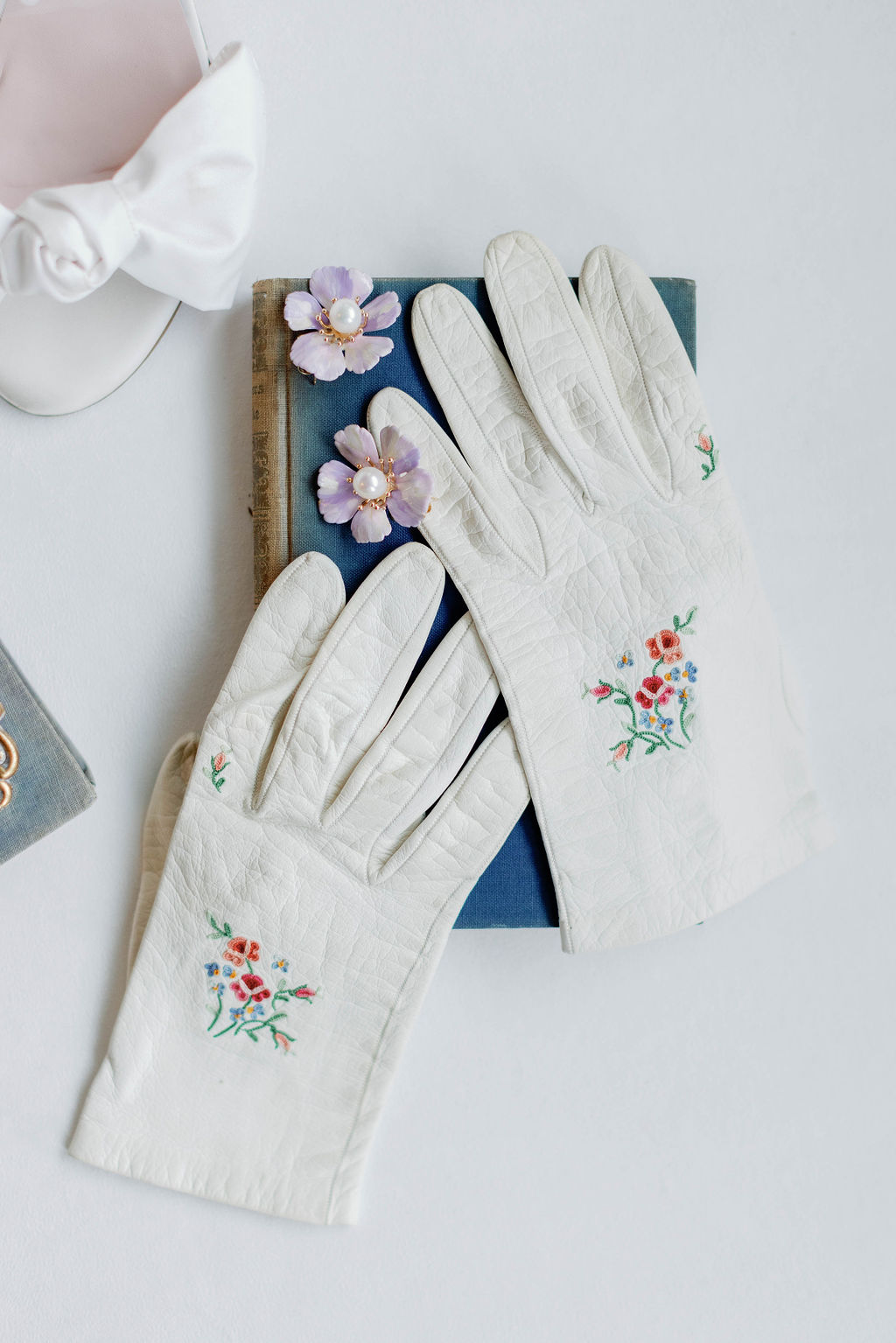 White Heirloom gloves with roses on them laid on top of an old vintage book along with a light purple floral earring with a pearl in the center taken at The Grand Lady Wedding Venue in Austin Texas. | Photo by Texas Wedding Photographer Sarah Block of Sarah Block Photography